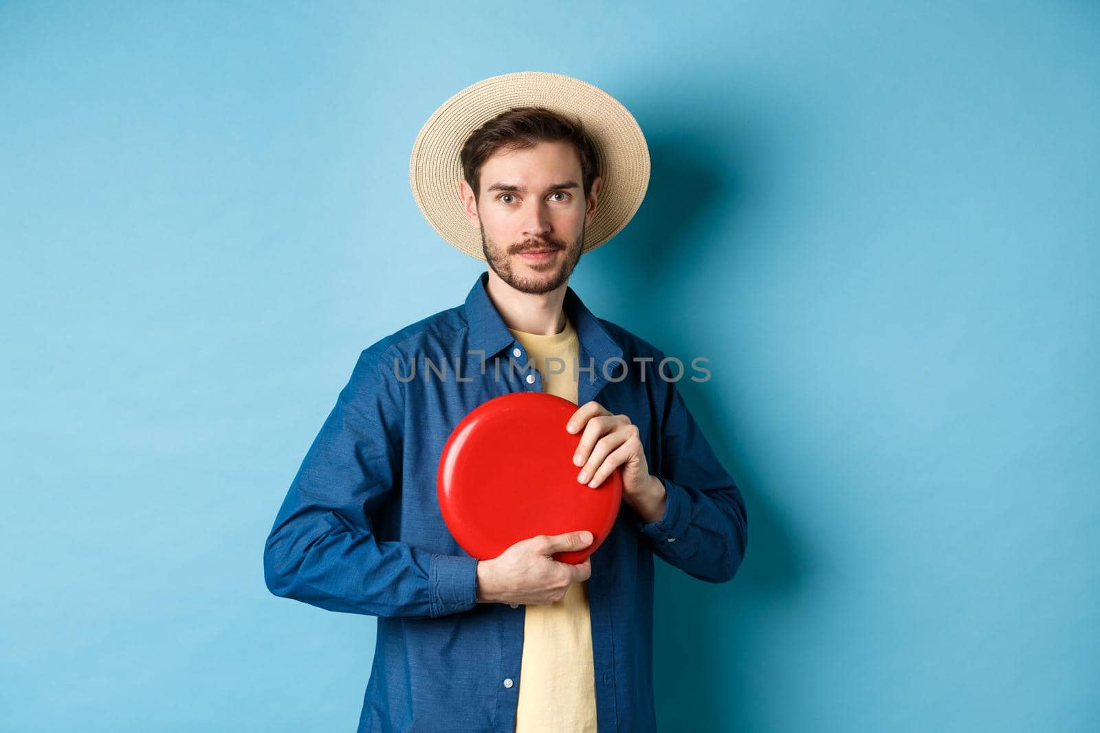Young man throwing frisbee, playing on summer vacation, standing in straw hat on blue background.