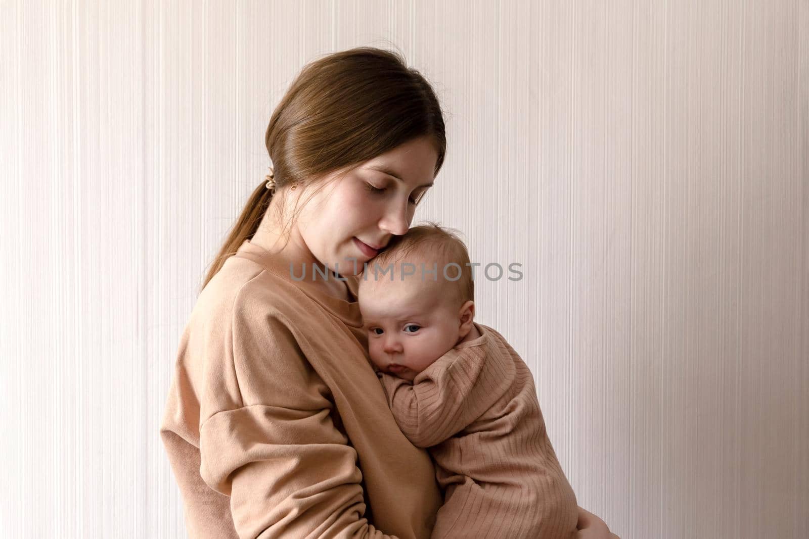 Cheerful beautiful young woman holding baby girl in her hands and looking at her with love at home.