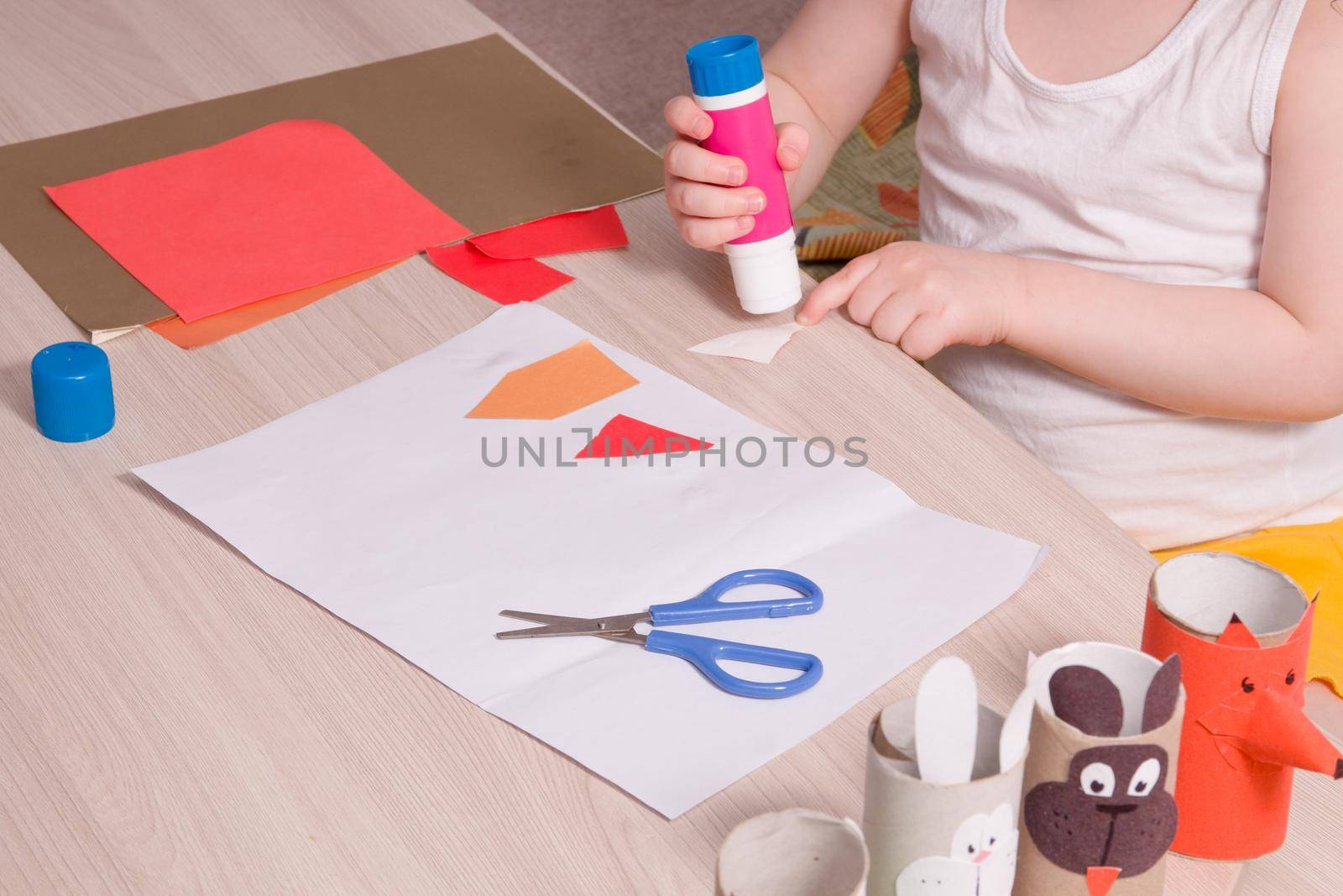 a small child smears paper with glue, children's safe scissors and colored paper on the table, what to do with the child at home, children's hands make an application
