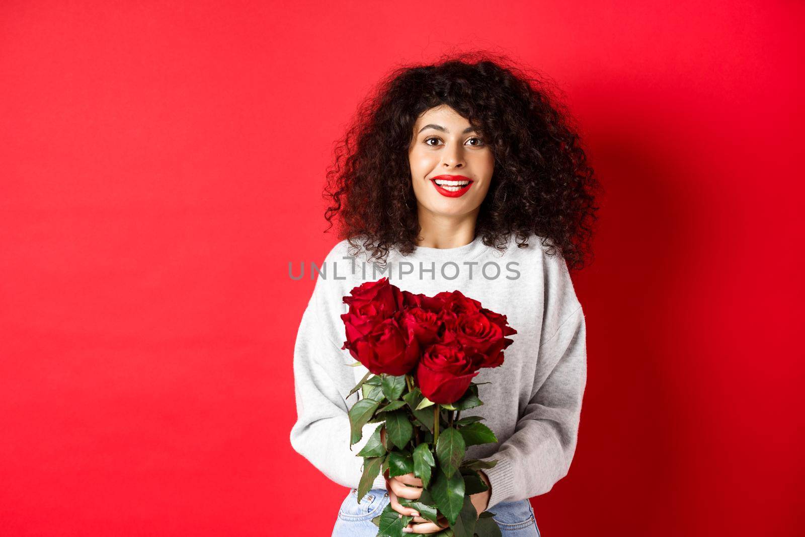 Romantic woman with curly hairstyle receive bouquet of red roses on Valentines day, looking tender at lover, standing on studio background.