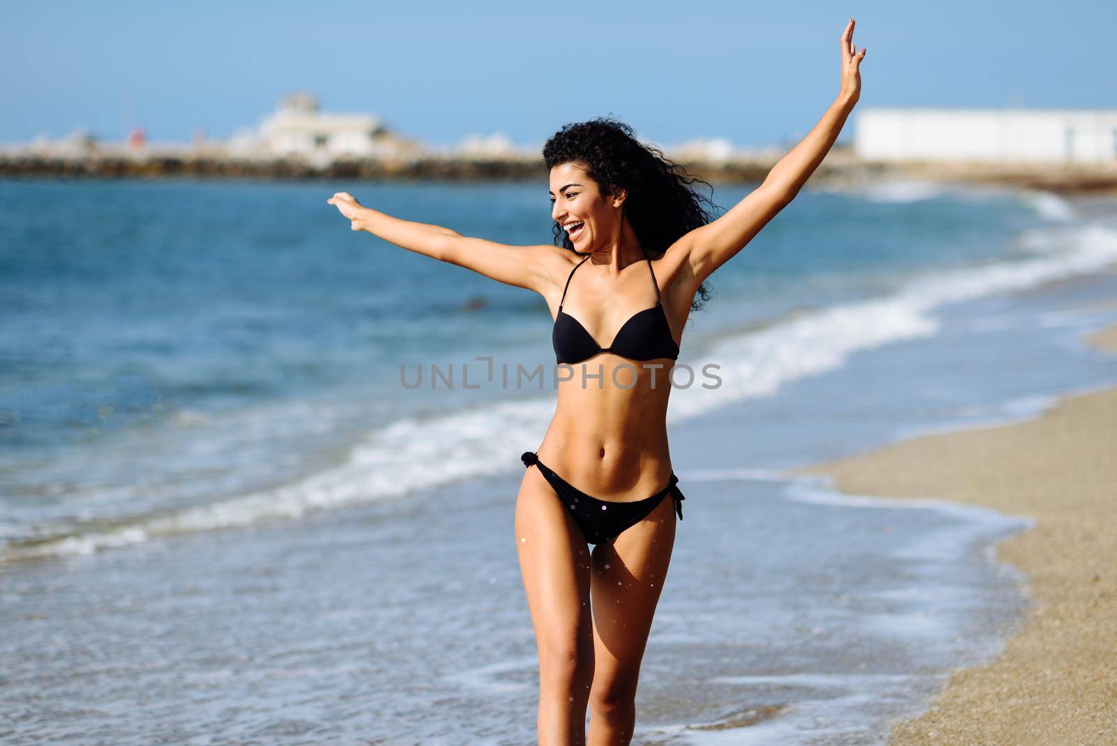 Young arabic woman with beautiful body in swimwear on a tropical beach with open arms. Brunette female smiling with curly long hairstyle wearing black bikini.