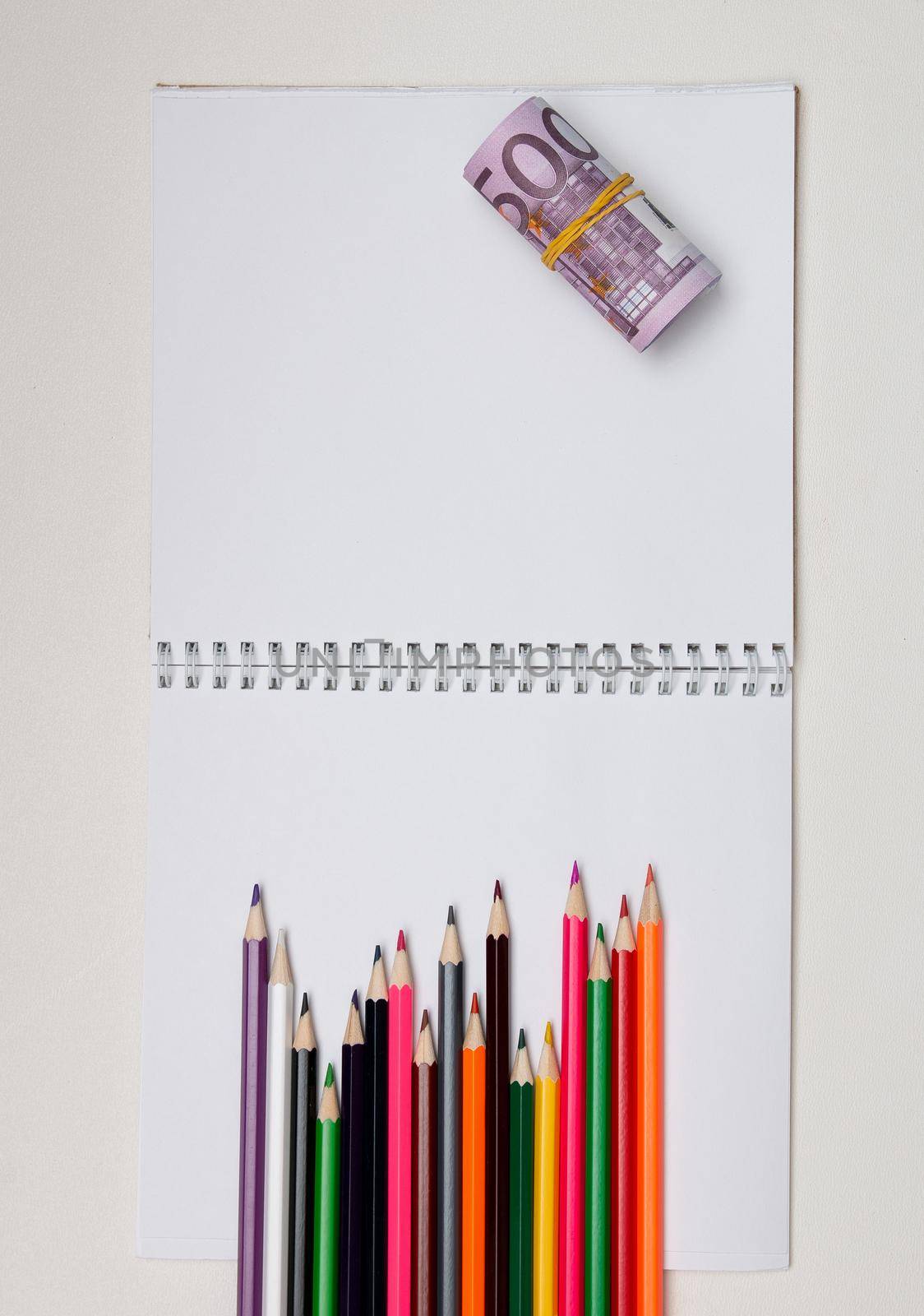 colored pencils lie on a white notebook with a spring and readable sheets of euro banknotes money on a white table pencil sharpening sharpen color top view spring copybook copy space