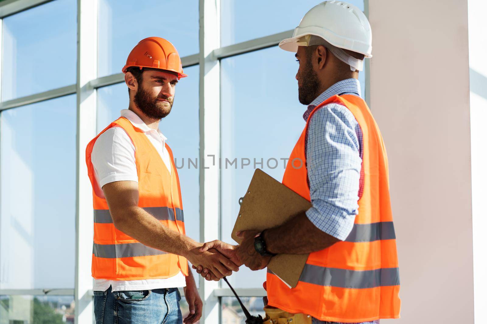 Two men engineers in workwear shaking hands against construction site. by Fabrikasimf