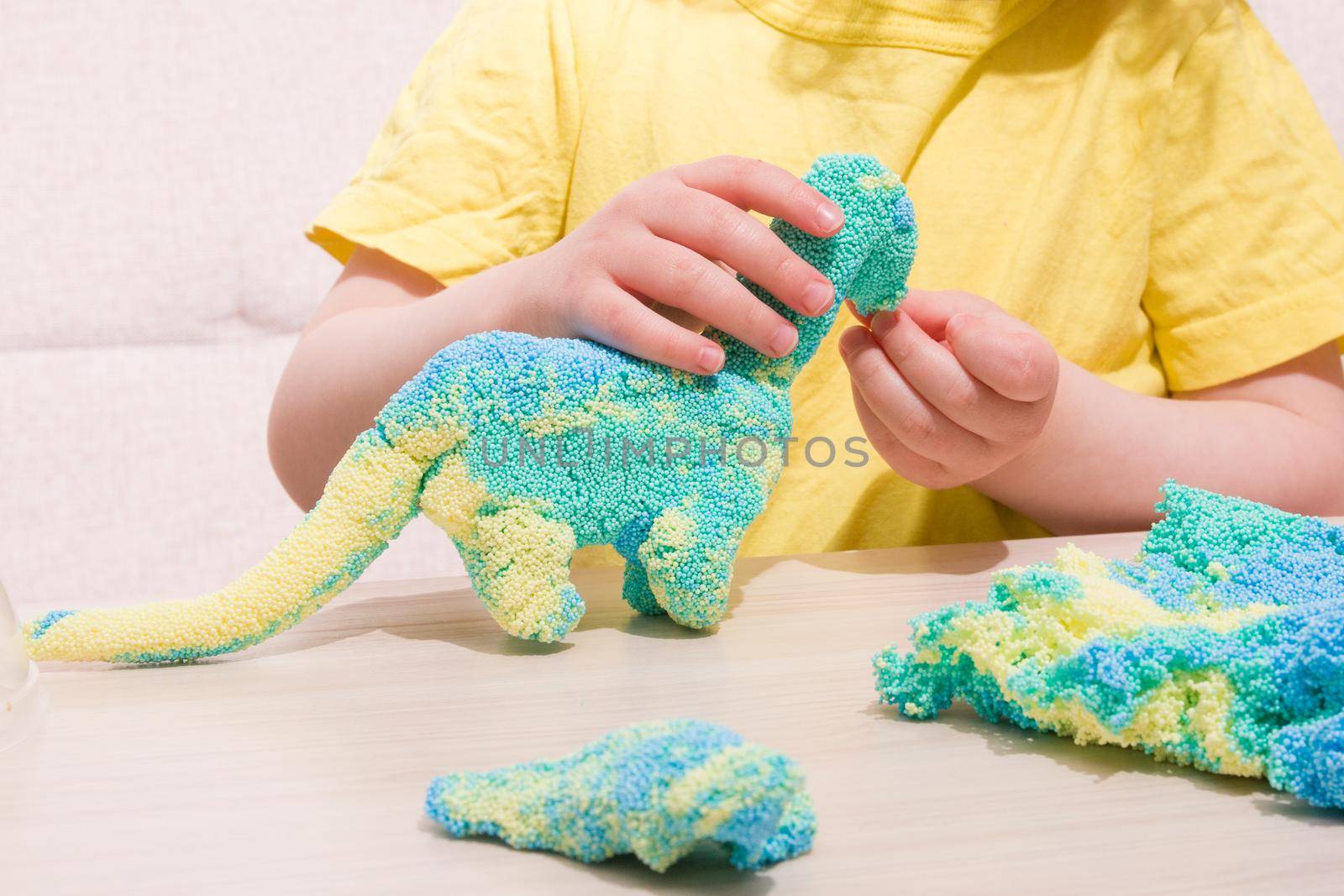 the child plays plasticine from polymer granules at the table, the original plasticine from tiny balls, the child plays with a dinosaur from plasticine, what to play with the child at home