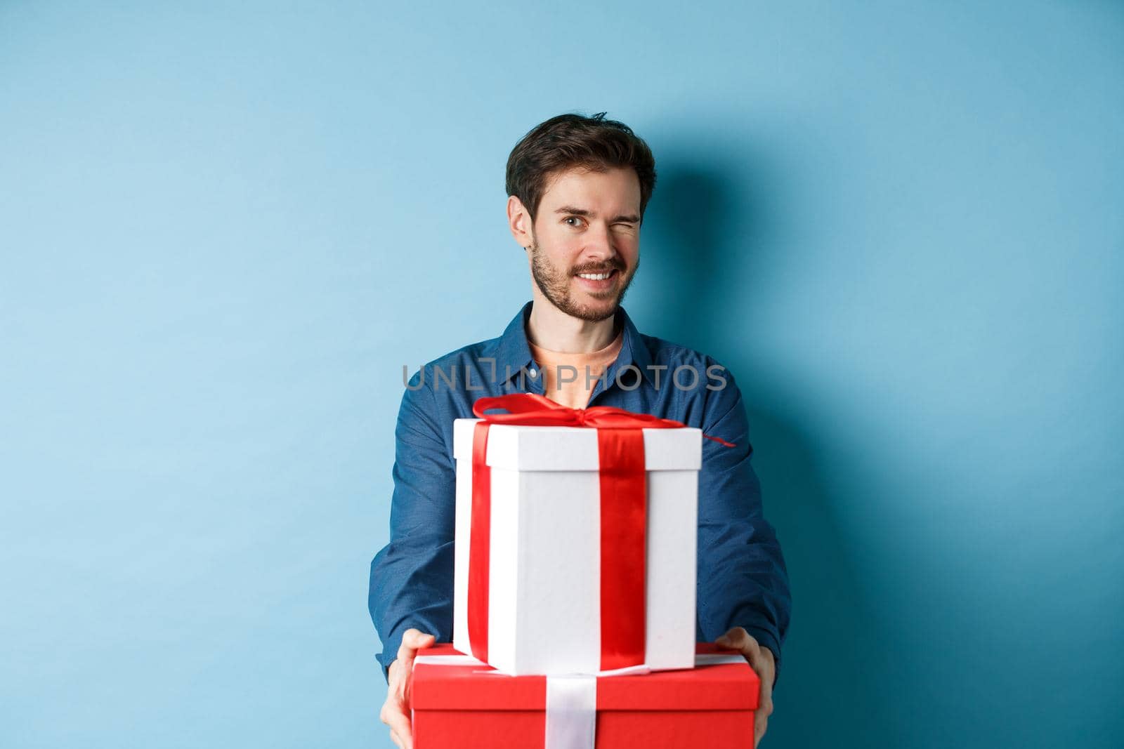 Handsome boyfriend winking at camera and wishing happy valentines day, extending hands with gift boxes, making romantic surprise, standing over blue background by Benzoix