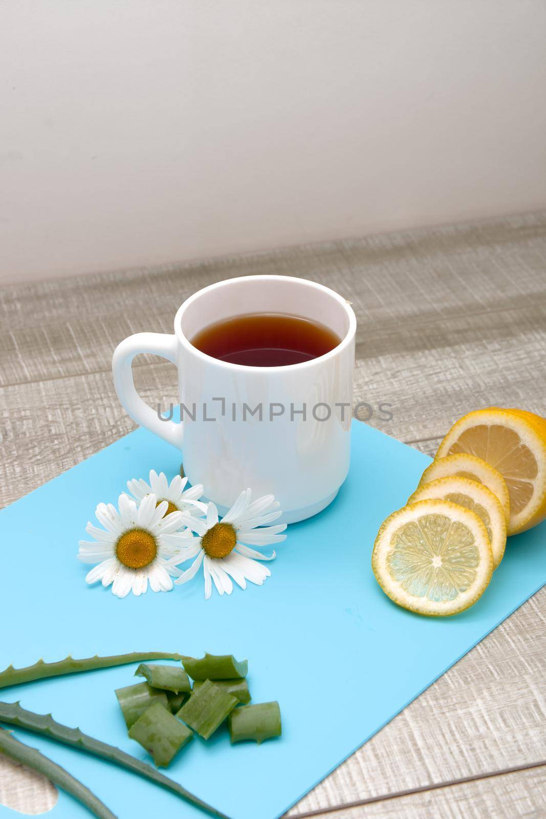 proper nutrition, white cup with chamomile tea on a blue cutting board with chamomile flowers, sliced lemon, aloe and polka dot fabric on a gray wooden table