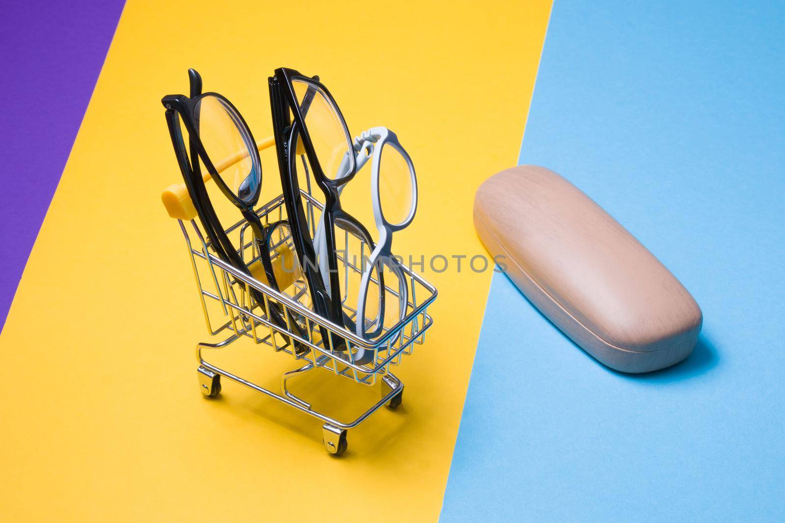 several different glasses in a small shopping trolley on a colorful background, a case for glasses on the background, a glasses store, children's glasses and glasses for adults