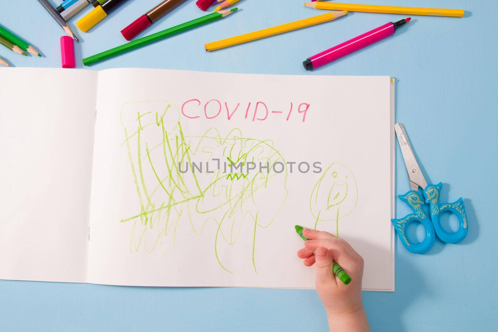 a small child draws green wax crayon in an album coronavirus, pencils on a blue background, top view, copy space, activities with children at home during quarantine