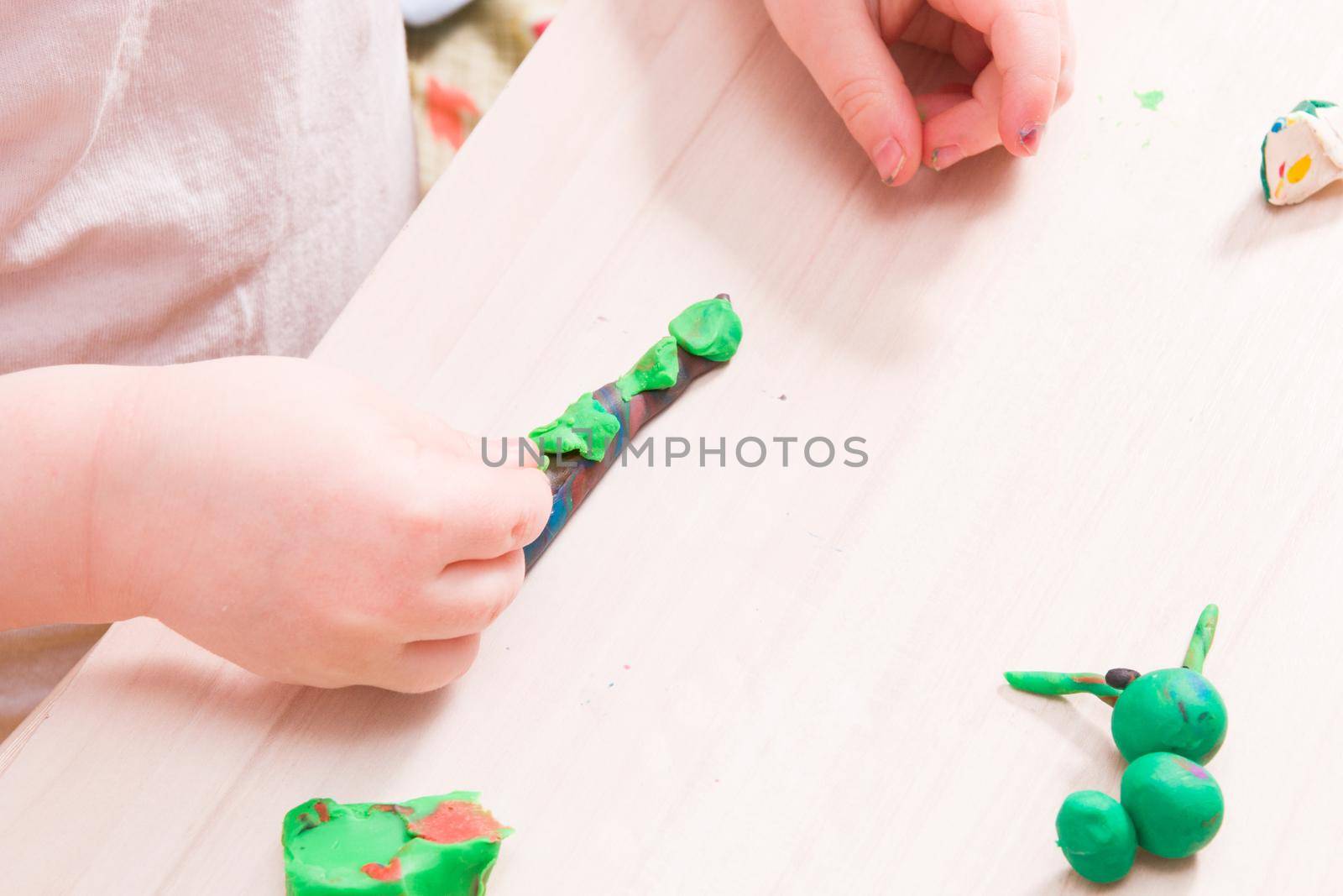 a small child sculpts a caterpillar from green plasticine on a wooden table, the development of fine motor skills of hands, playing with a child at home, a caterpillar from plasticine in the foreground by natashko