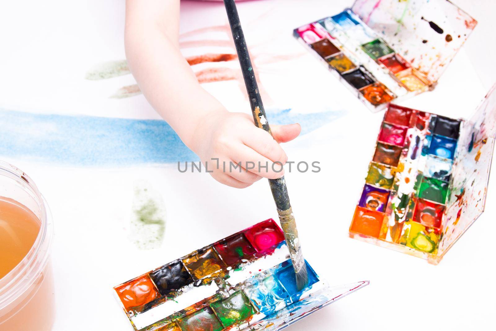the child paints with a brush watercolors on a white sheet, a table, a copy space, several packages of watercolor, a large art brush in his hand, a can of water by natashko