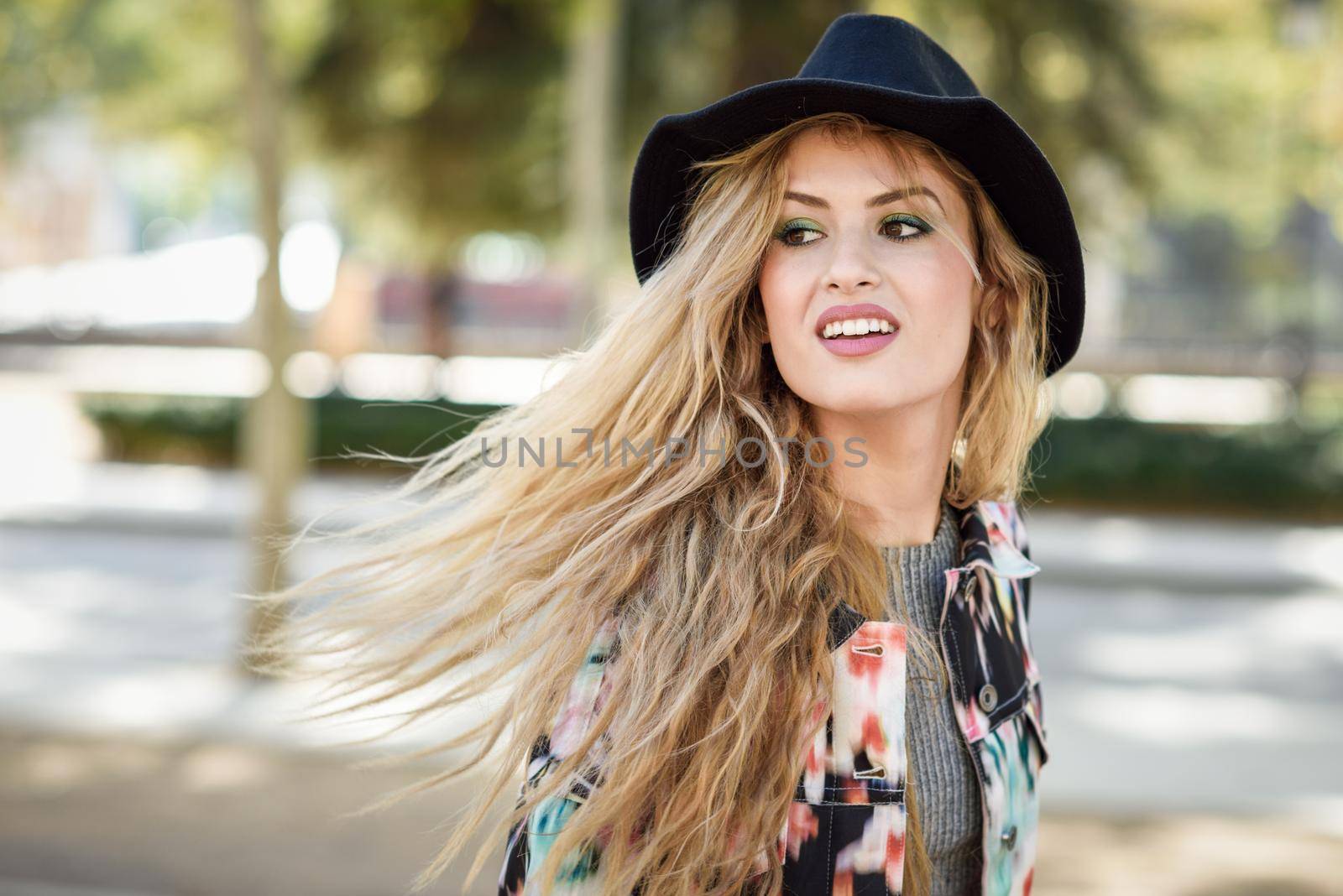 Young woman with flying hair wearing jacket and hat, happy in urban background