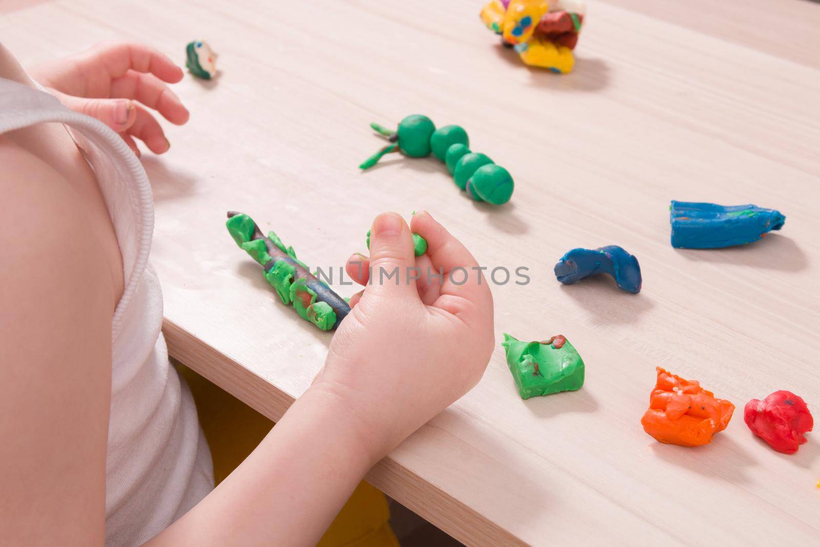 a small child sculpts a caterpillar from green plasticine on a wooden table, the development of fine motor skills of hands, playing with a child at home, a caterpillar from plasticine in the foreground by natashko