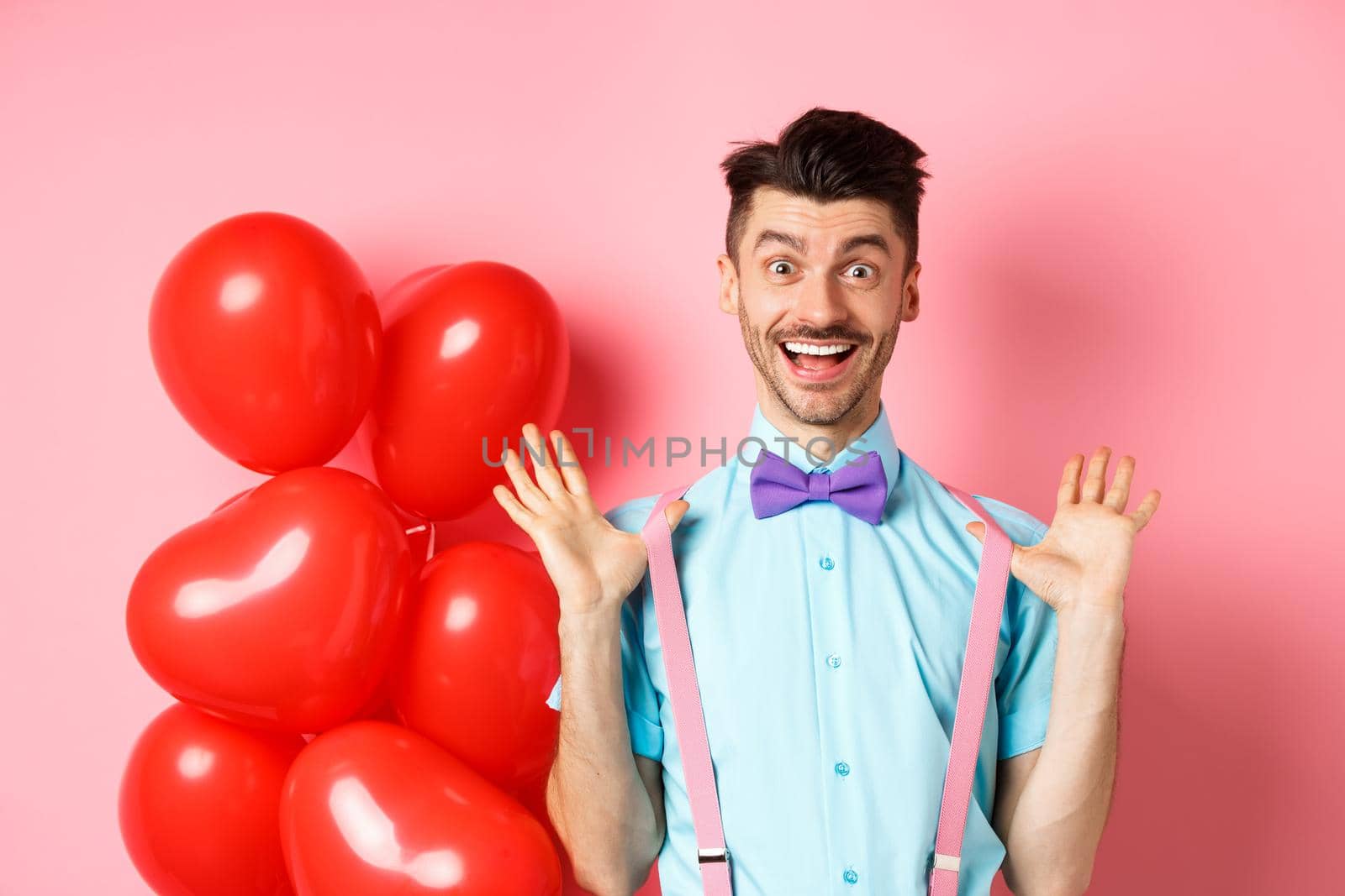 Valentines day concept. Happy young man looking surprised, raising hands up joyful, celebrating near big red hearts and pink background by Benzoix