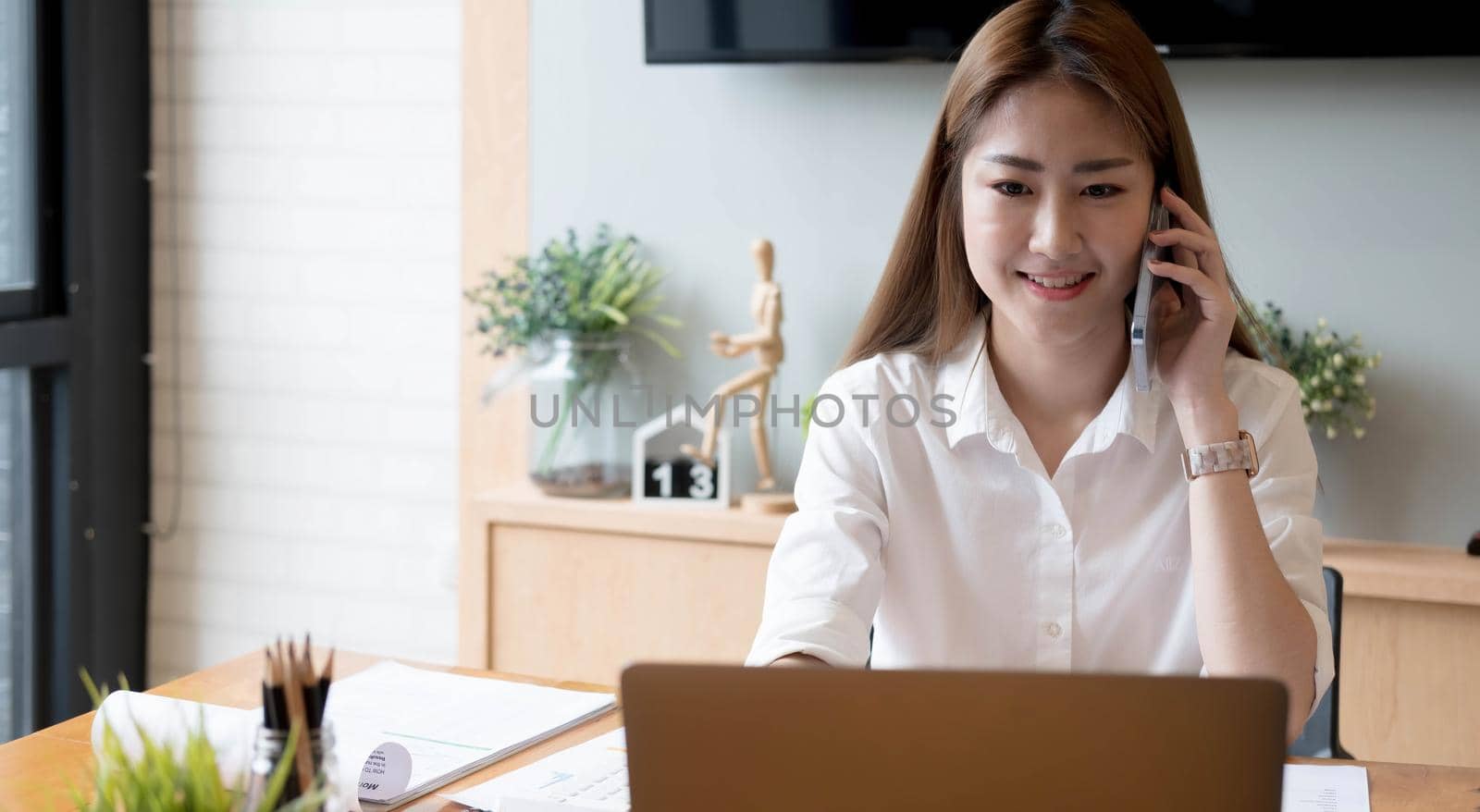 Smiling asian woman taking call on mobile phone during working on laptop computer for financial accounting number, paperwork statistics monthly report by wichayada