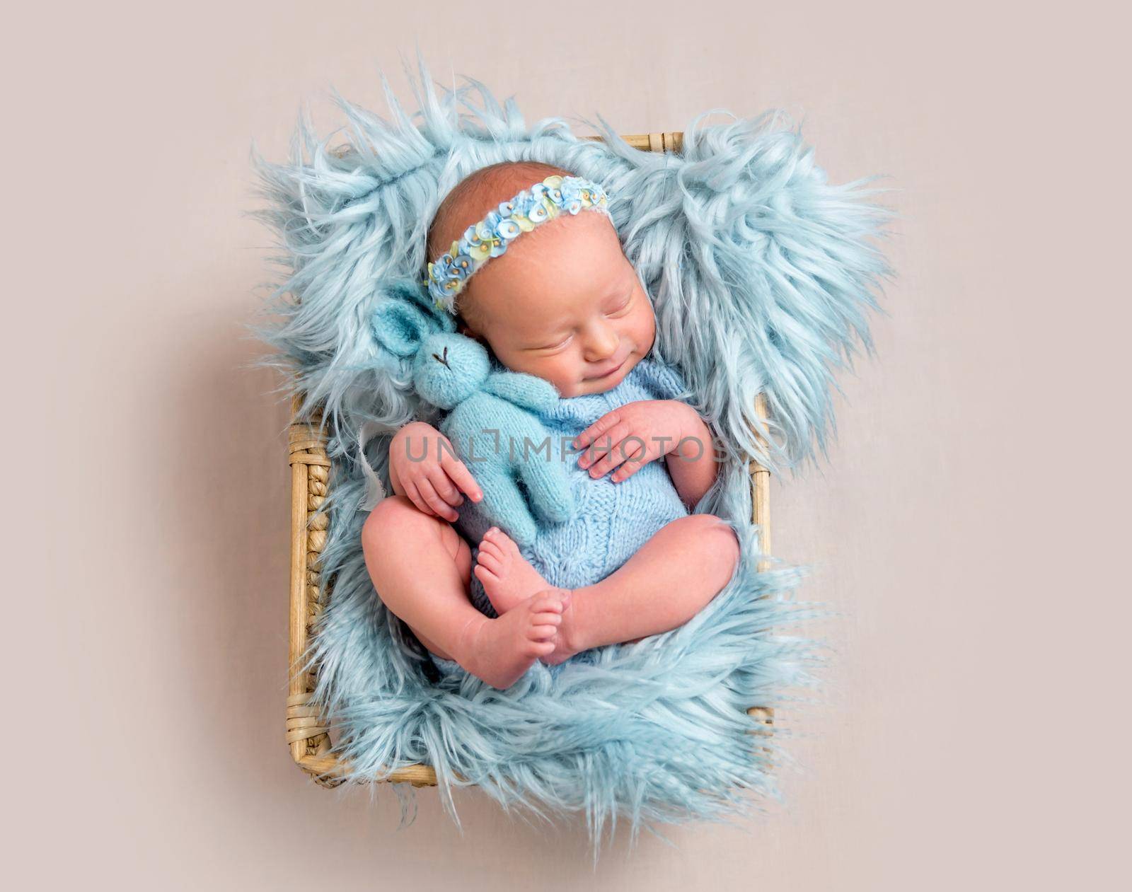 Happy newborn girl lying on her back and cuddling her toy. Little girl wearing blue headband and blue bodysuit lying on blue flokati rug and smiles in sleep while hugging her hare toy