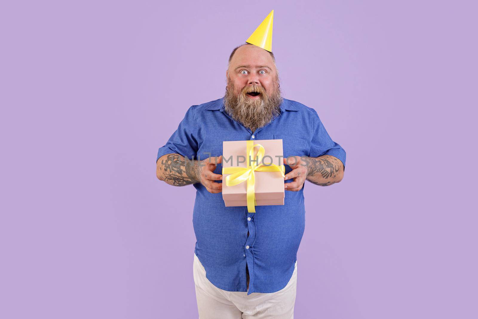 Excited mature plump man wearing tight blue shirt and party hat holds present posing on purple background in studio