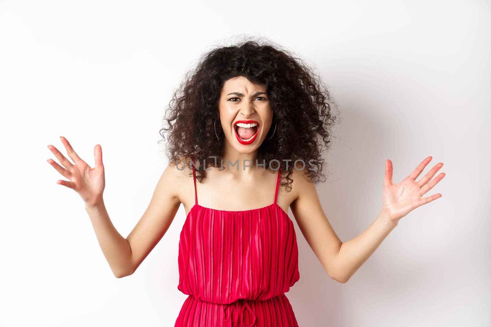 Angry young woman with curly hair, wearing red dress, screaming and having an argument, looking with hatred and anger, standing over white background by Benzoix