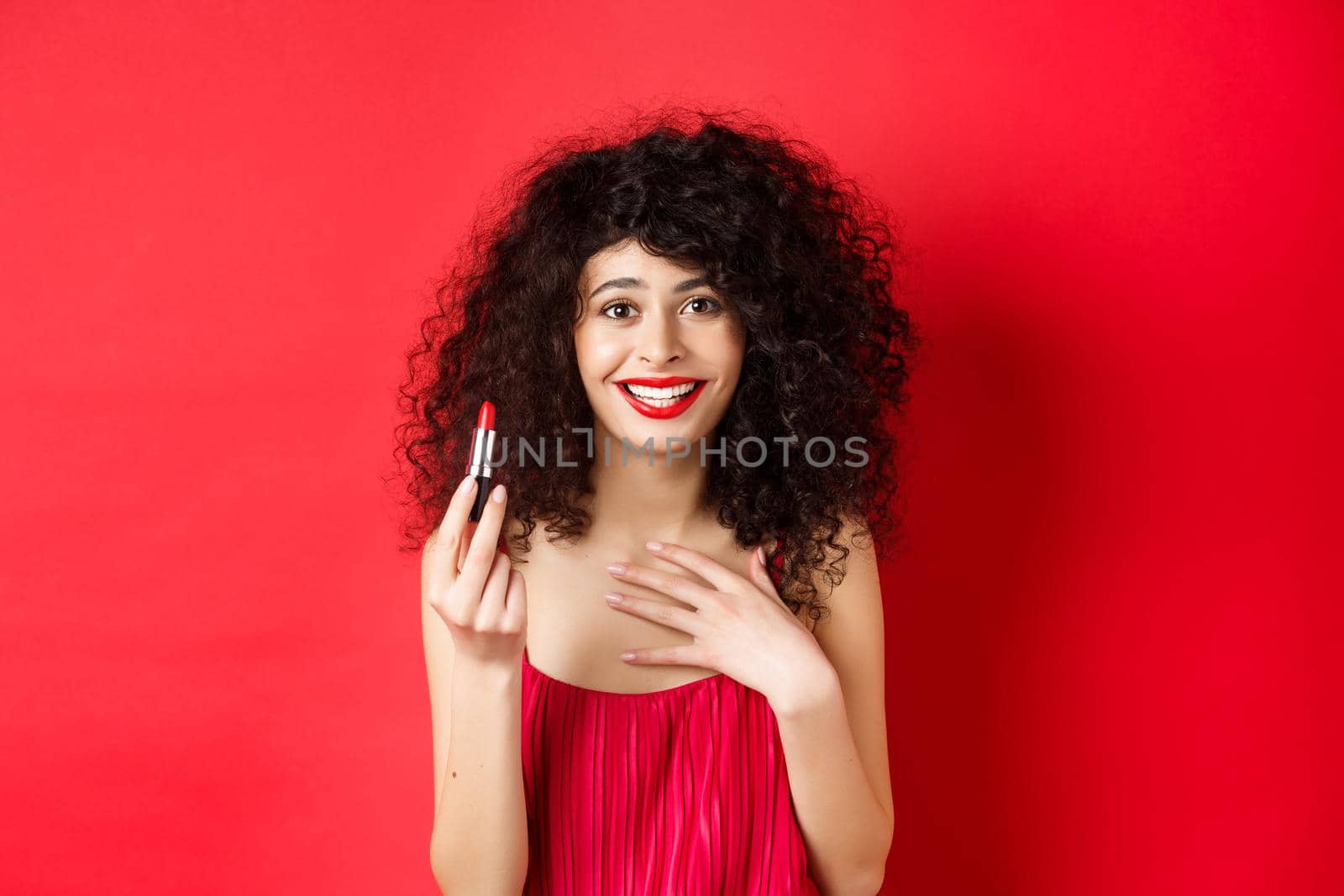Beauty and make up concept. Beautiful female model with curly hair, wearing evening dress, showing red lipstick and smiling, standing on white background by Benzoix