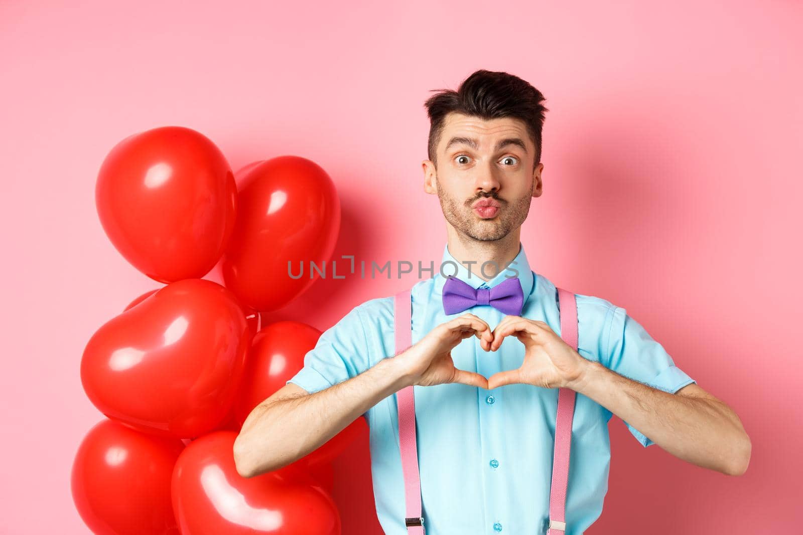 Valentines day concept. Cute boyfriend pucker lips for kiss, standing in bow-tie with heart gesture, say I love you, standing on pink background.