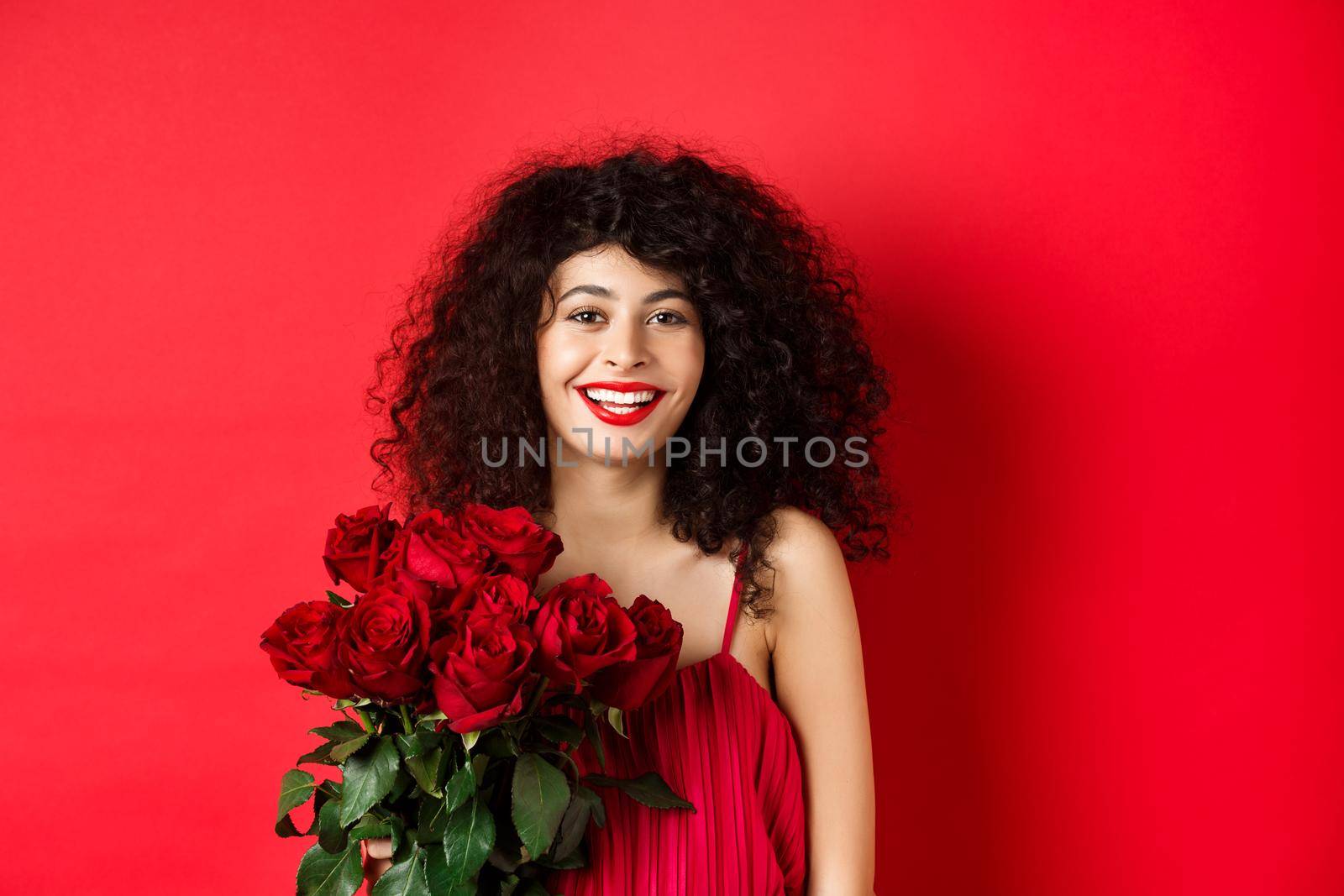 Happy fashionable female model with bouquet of red roses, smiling and looking cheerful at camera, studio background.
