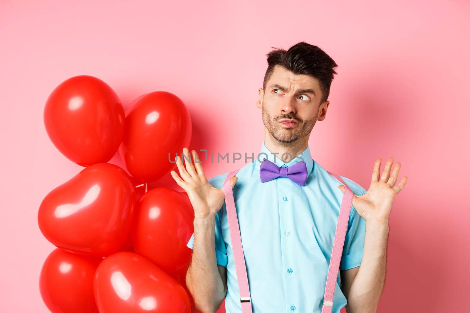 Valentines day concept. Pensive young man in bow-tie, raising hands up and looking left while thinking, making decision, standing on romantic pink background and heart balloons by Benzoix