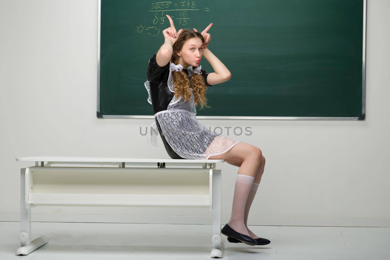 Joyful happy girl student sitting at the desk. Smiling pretty shoolgirl with braided in two plaits hair wearing retro soviet school uniform sitting on desk in front of blackboard