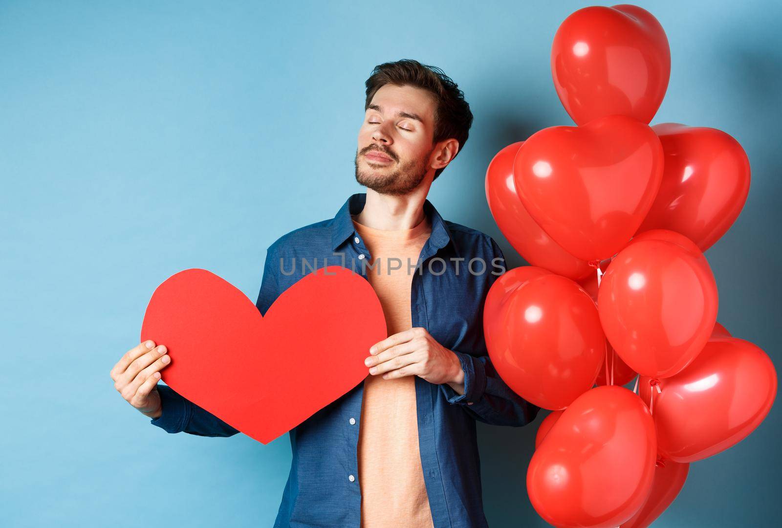 Valentines day and love concept. Dreamy man with closed eyes, holding romantic red heart cutout and standing near hearts balloons, blue background by Benzoix