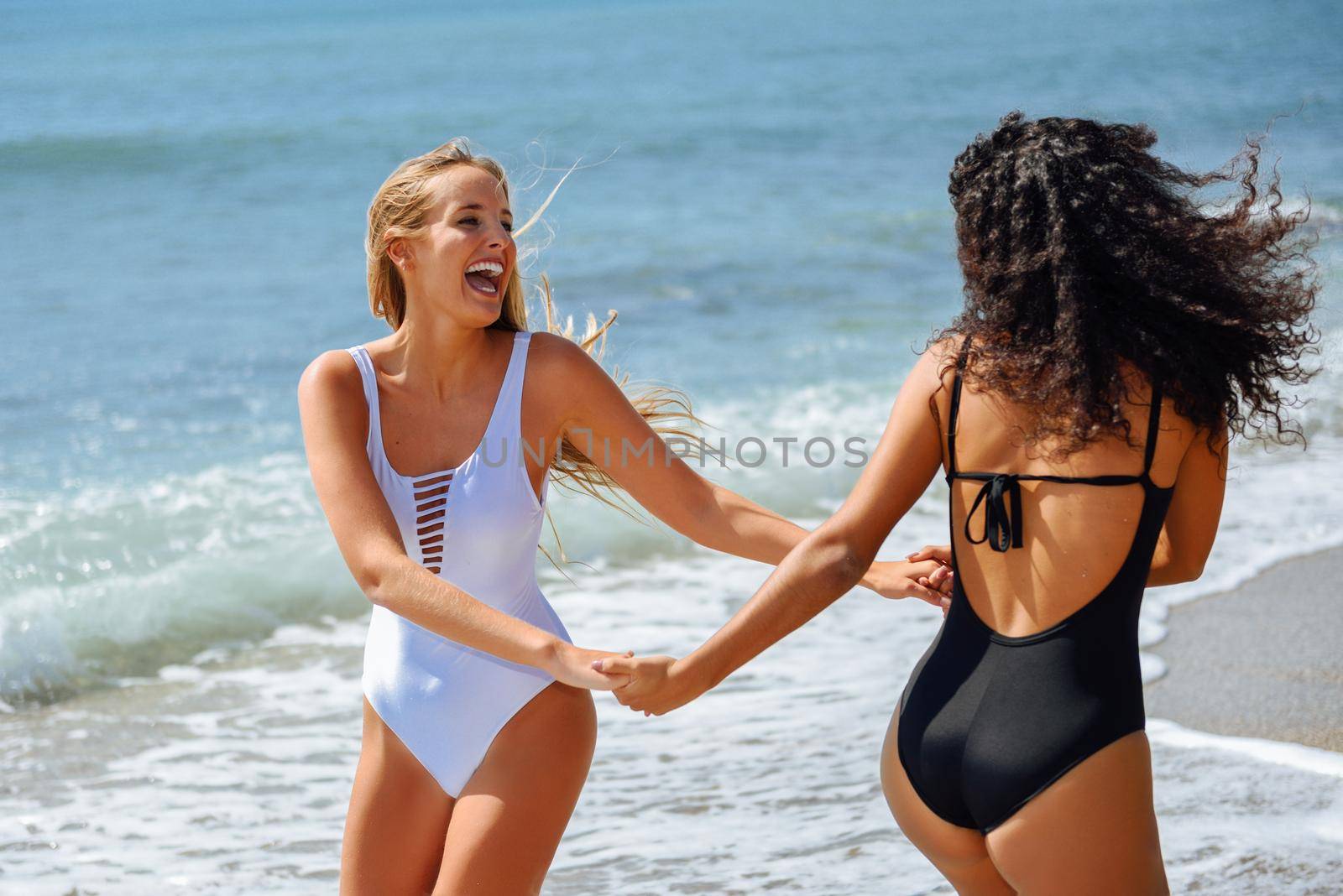Two young women with beautiful bodies in swimwear having fun with their hands caught on the beach. Funny caucasian and arabic females wearing black and white swimsuits