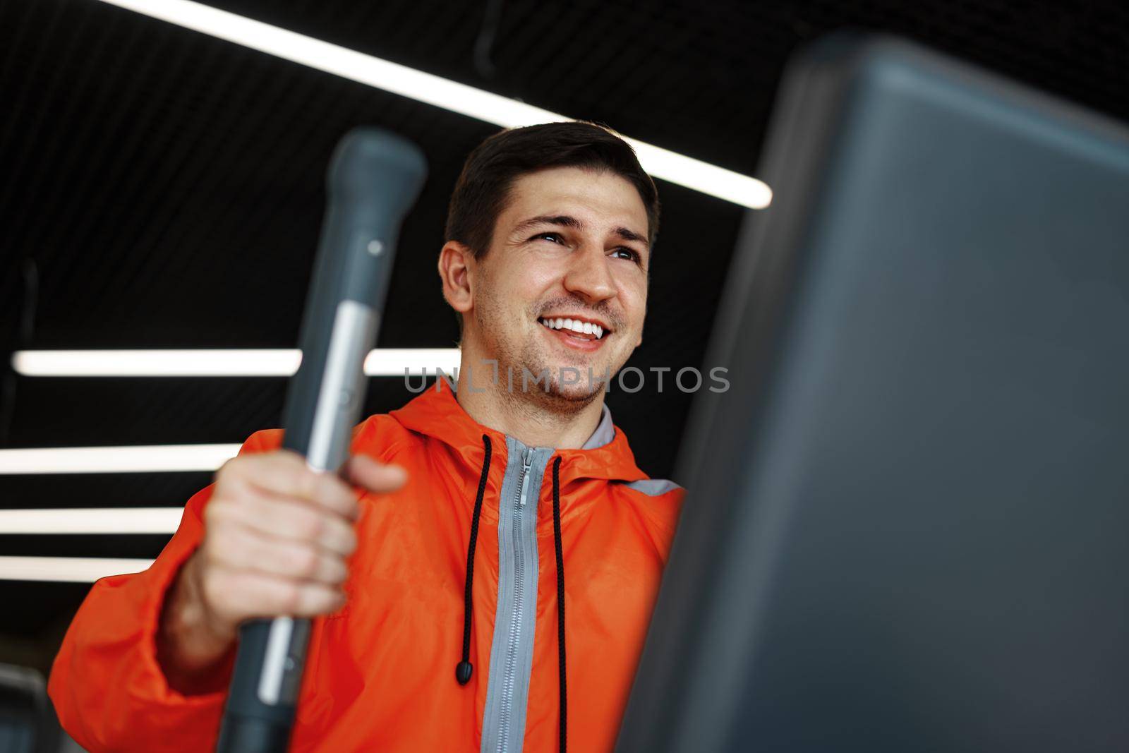 Portrait of a young man in orange windbreaker workout on a fitness machine at a gym.