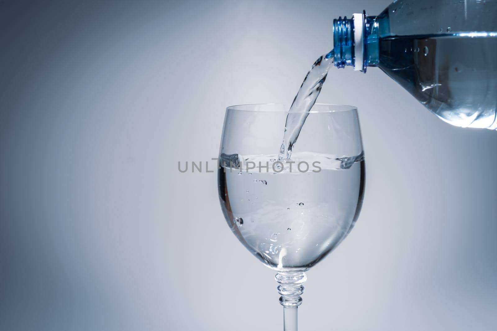 Pouring water into a glass goblet from a plastic bottle. Light contrasting background with veneering.