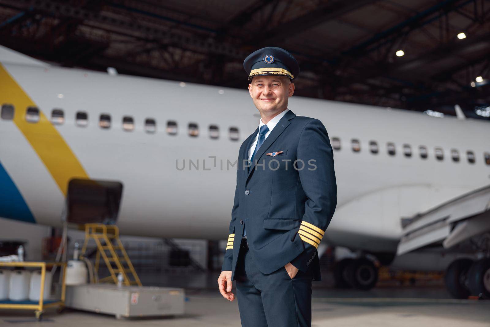 Portrait of smiling pilot in uniform looking at camera, standing in front of big passenger airplane in airport hangar. Aircraft, occupation, transportation concept