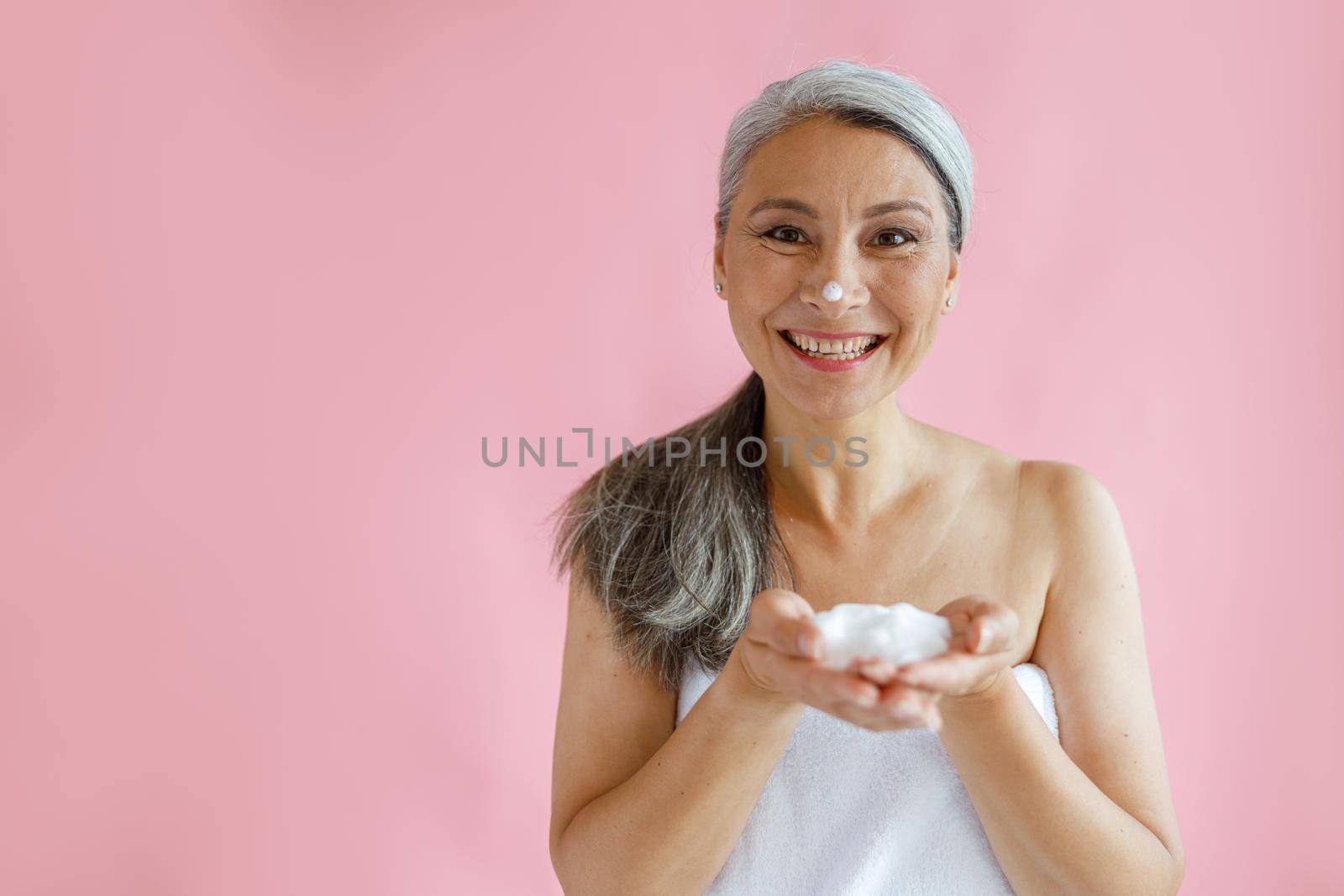 Jouful silver haired Asian lady with handful of foam stands on pink background by Yaroslav_astakhov