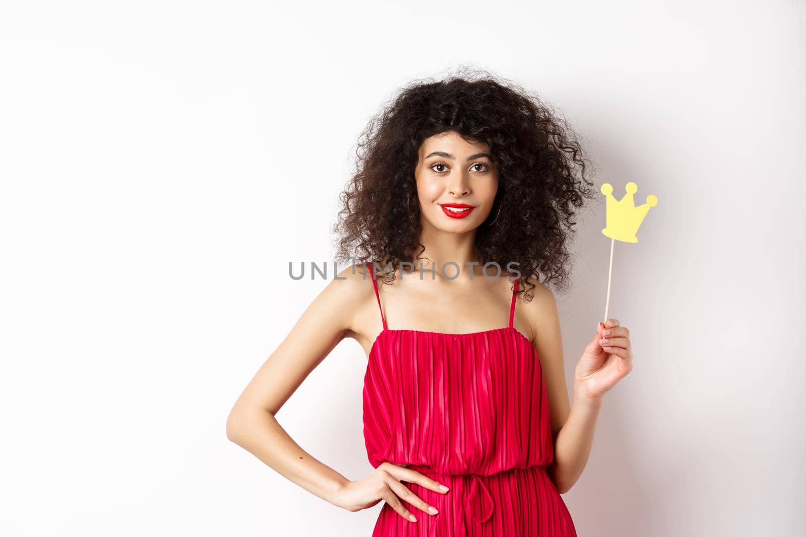 Stylish young woman in red dress, feeling confident and sassy, holding crown and smiling, standing over white background.