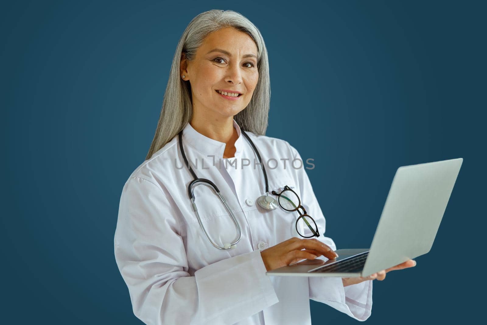 Smiling mature woman doctor in white coat works on laptop standing on blue background by Yaroslav_astakhov