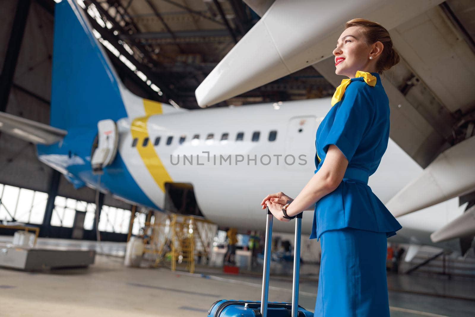 Pretty air stewardesses in bright blue uniform smiling away while standing with suitcase in front of passenger aircraft by Yaroslav_astakhov