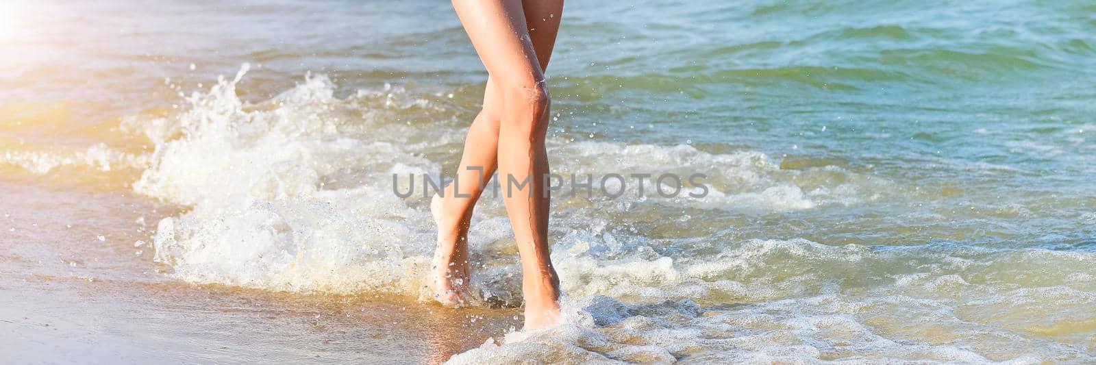 Running girl legs on the sea beach. Long summer banner with copy space. Close up of a young girl's legs walking or running on the beach with waves and seafoam.