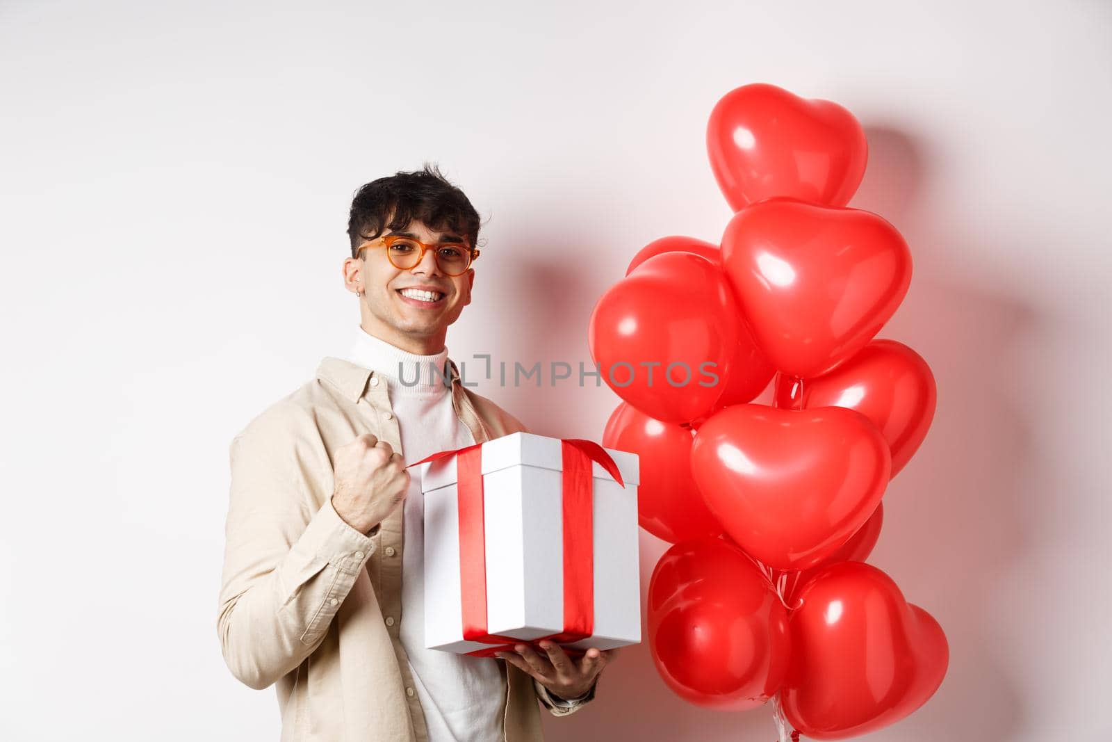 Valentines day and romance concept. Happy and confident boyfriend prepare gift for lover, saying yes and smiling, holding romantic gift, standing near red hearts balloons.