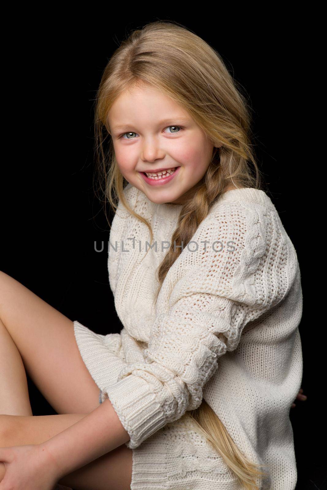 Close up portrait of beautiful girl with braid. Smiling cute little girl wearing warm knitted jumper sitting on floor against black background in studio