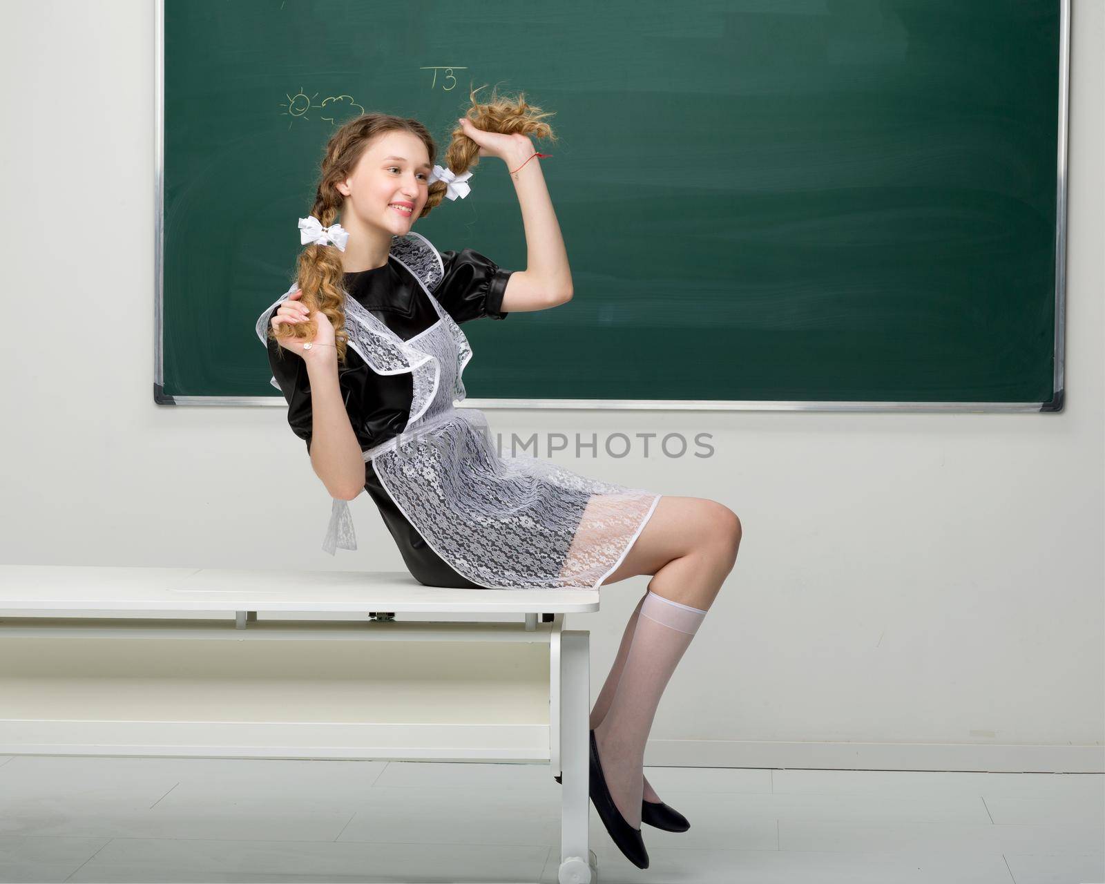 Joyful girl student holding her two long braids. Smiling pretty shoolgirl with braided in two plaits hair wearing retro soviet school uniform sitting on desk in front of blackboard