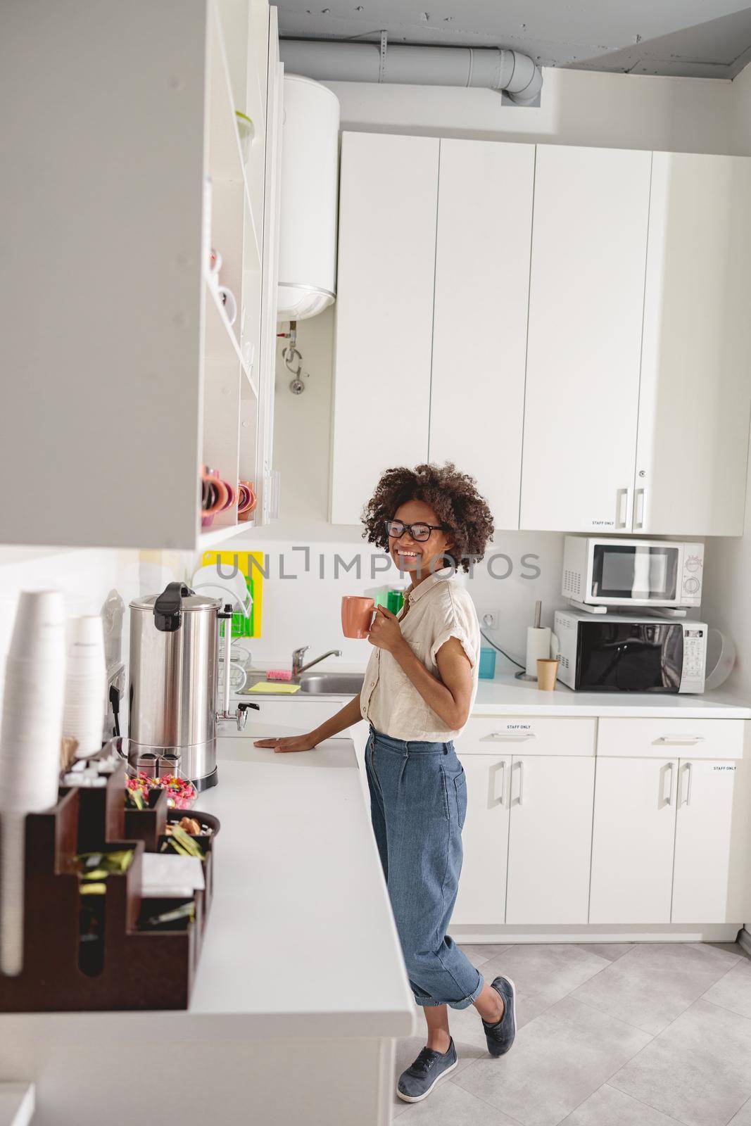 Full-length view of Afro American lady standing on office kitchen while holding cup with hot drink