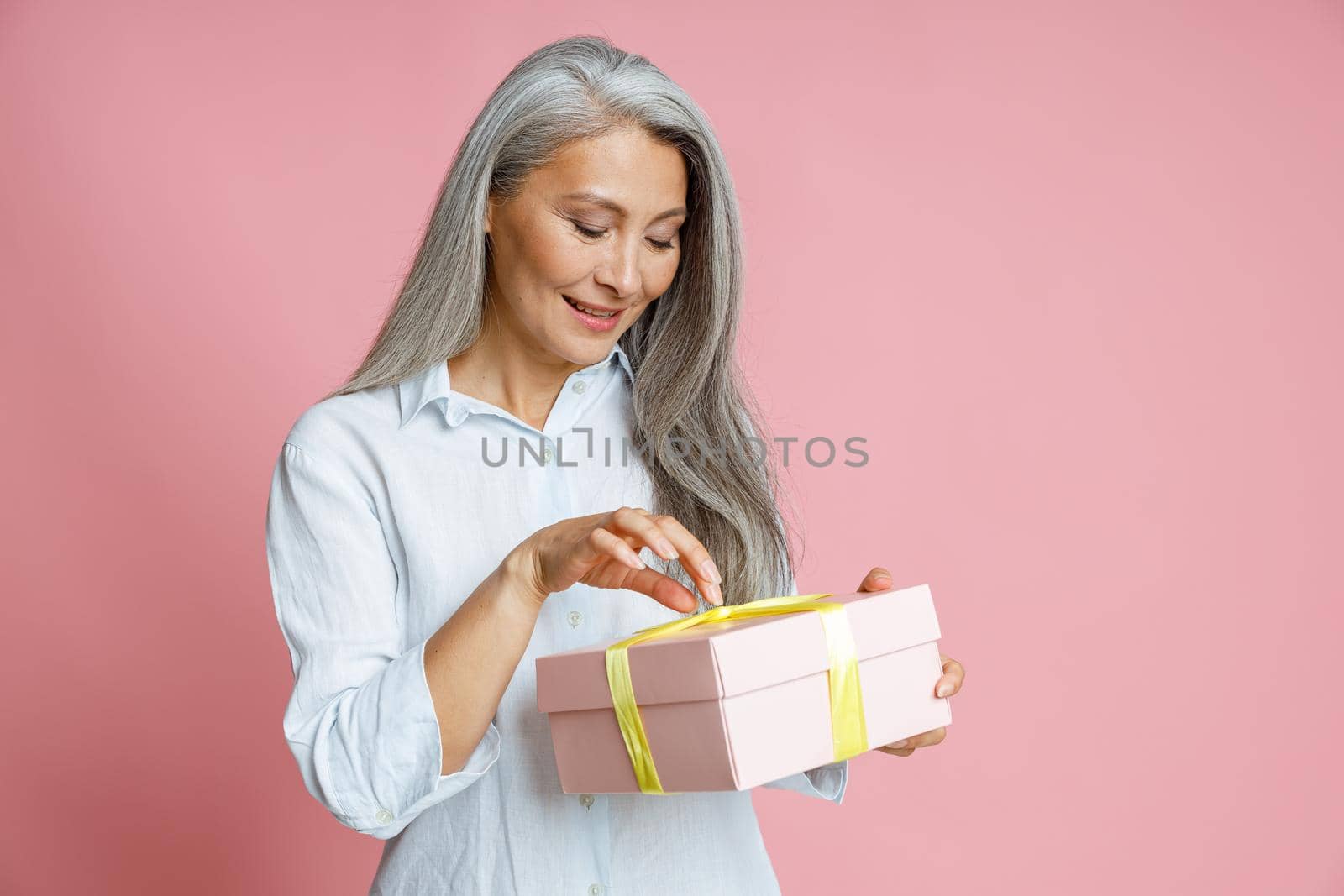 Smiling middle aged Asian woman with long grey hair opens gift box decorated with ribbon posing on pink background in studio