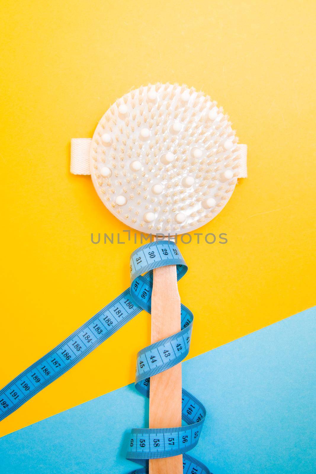 wooden brush for dry body massage on a blue and yellow background, measuring tape wrapped around the brush handle, copy space, peeling and body care at home