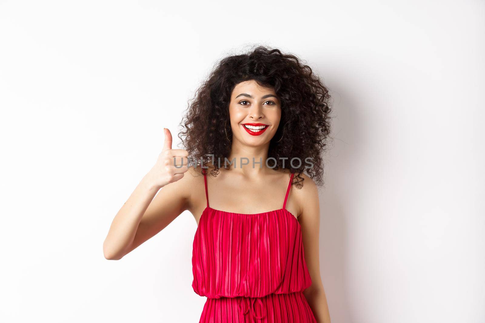 Cheerful woman with red lips and elegant dress, showing thumb up and smiling, recommending product, standing over white background.