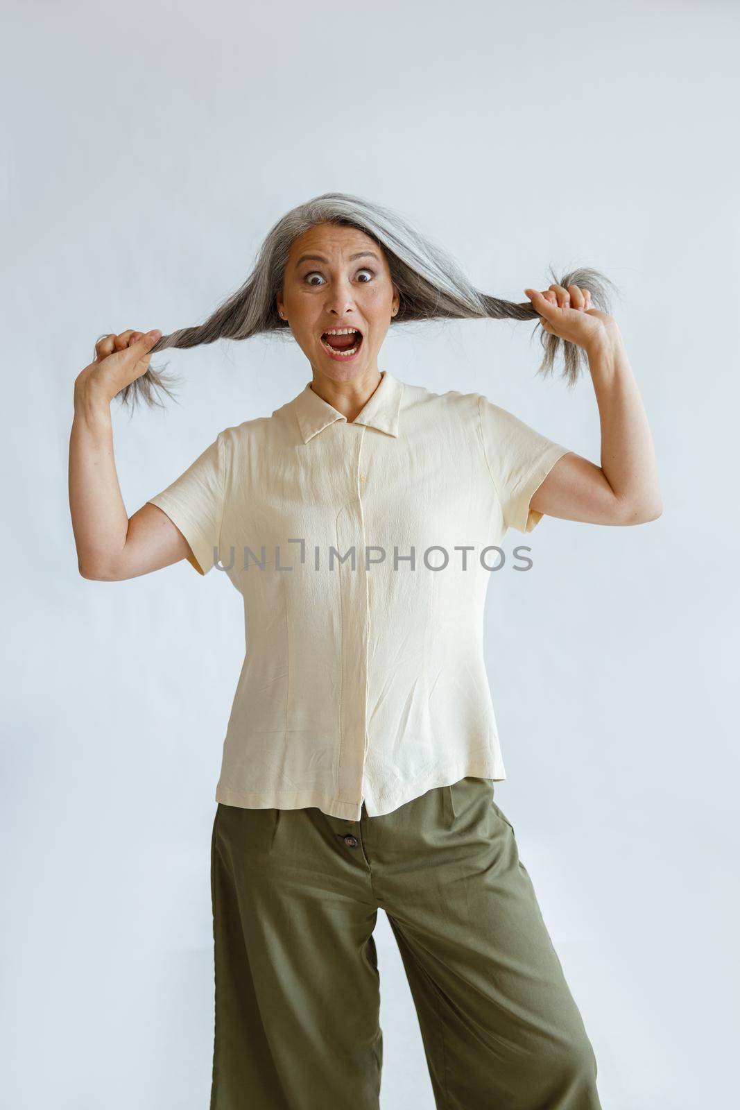 Shocked Asian woman holds hoary hair locks on light grey background by Yaroslav_astakhov
