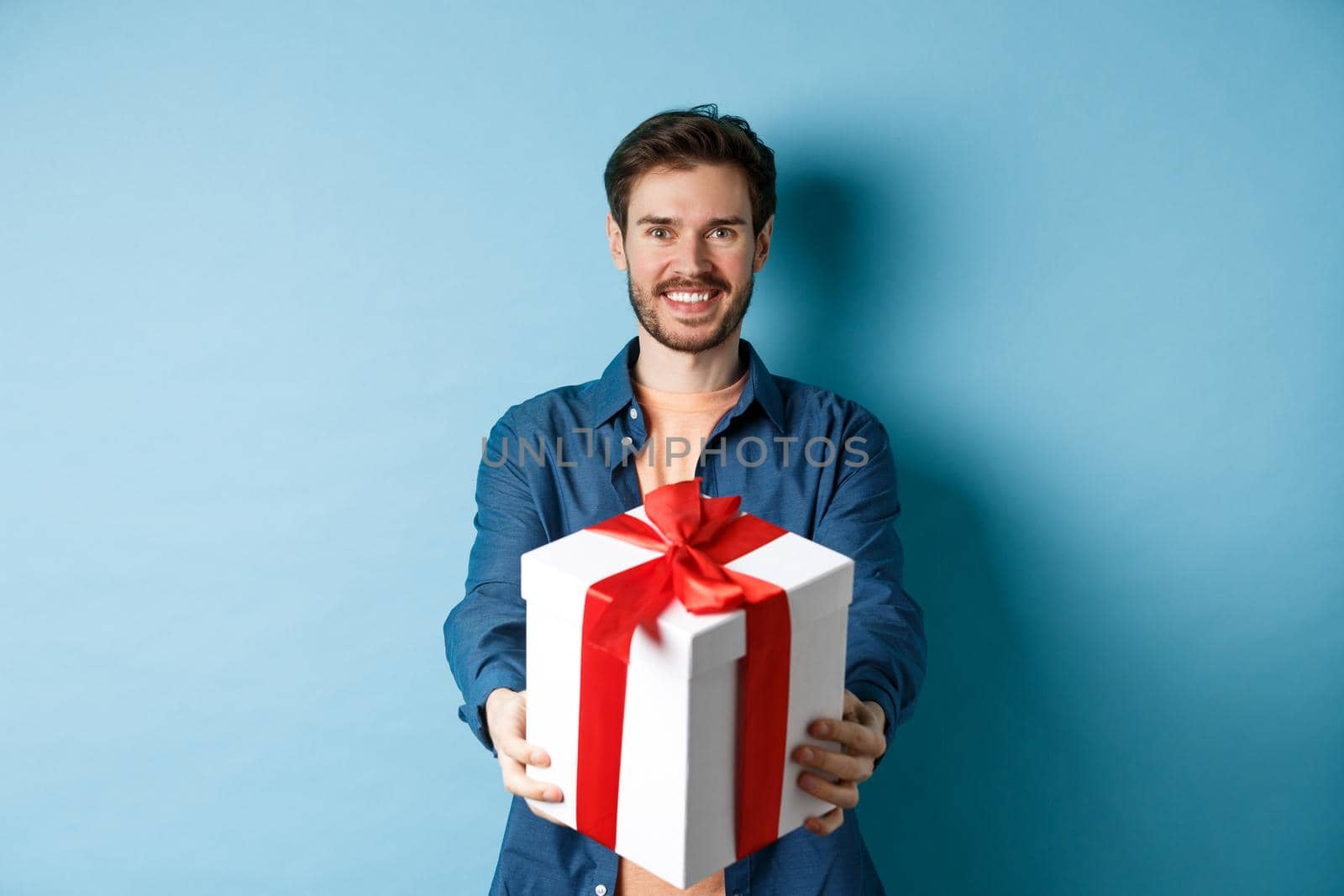 Valentines day. Handsome smiling man extending hands with gift box, wishing happy holiday. Guy making surprise present and looking cheerful, standing over blue background.