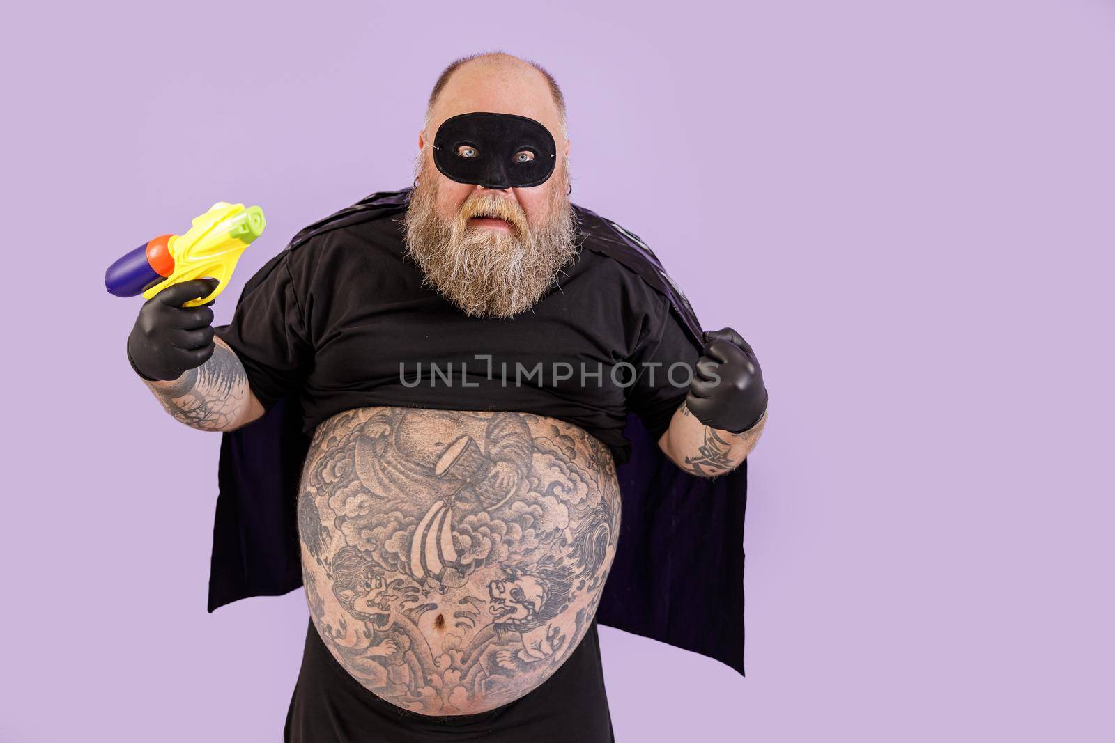 Funny evil obese man in carnival costume holds toy blaster posing on purple background by Yaroslav_astakhov