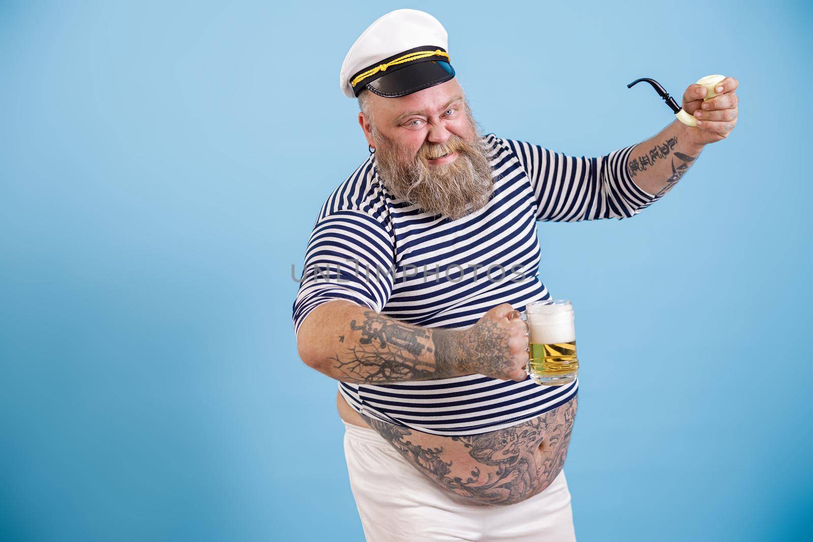 Grinning mature plus size man with beard in sailor costume holds smoking pipe and glass of tasty beer on light blue background in studio