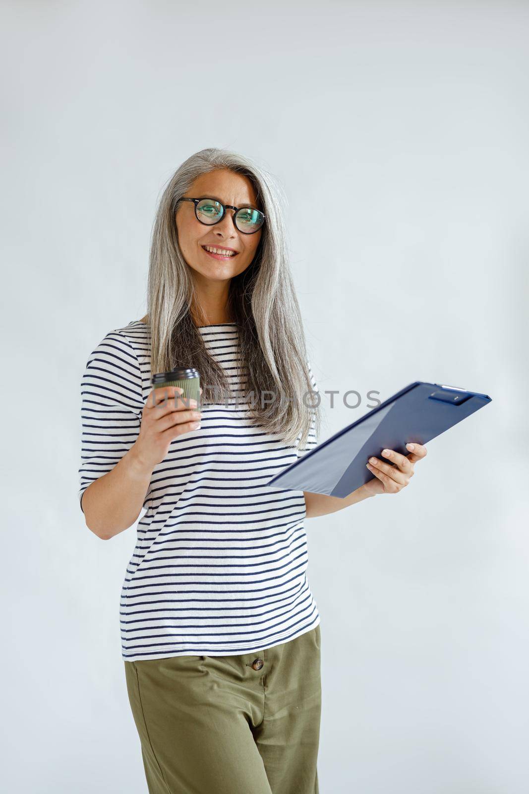 Beautiful mature Asian woman in casual clothes holds clipboard and cup standing on light grey background in studio