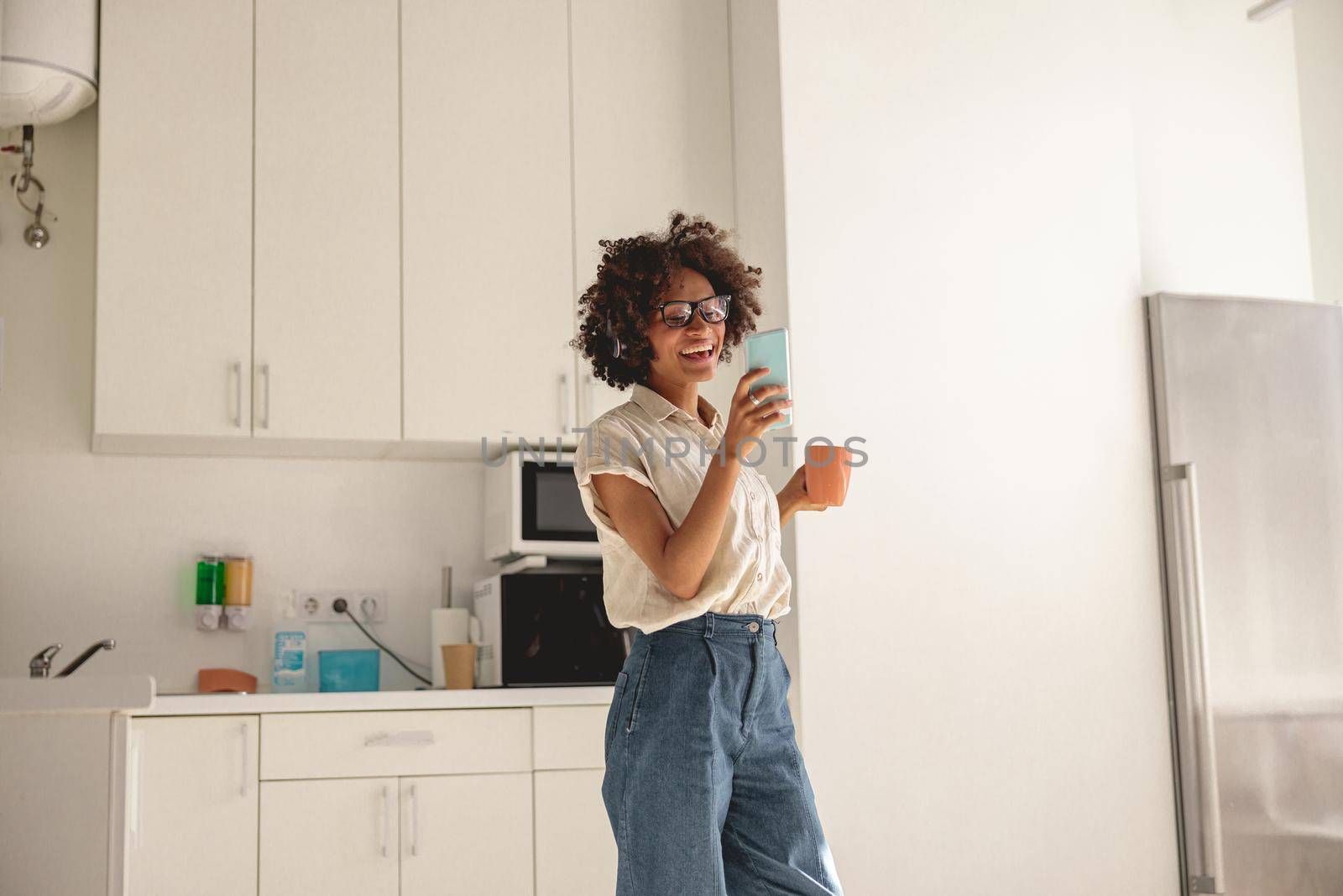 Happy Afro American woman holding phone and cup on the kitchen by Yaroslav_astakhov