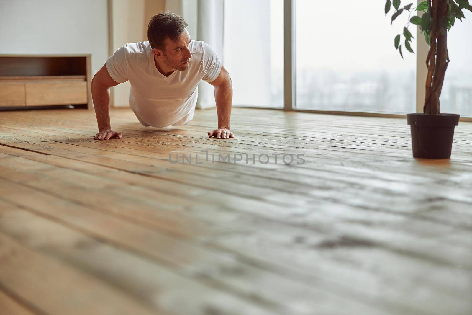 Low angle of athletic male doing push ups near window during domestic workout during quarantine